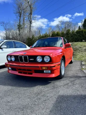 BMW M3 E30 rouge, vue de face trois quarts, calandre à double haricot et quatre phares ronds.