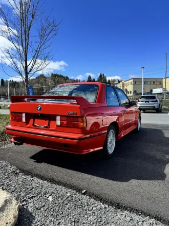 BMW M3 E30 rouge, trois quarts arrière, aileron M3 visible.
