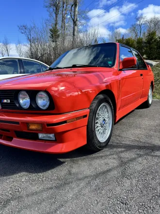 BMW M3 E30 rouge, trois quarts avant, jantes BBS visibles.