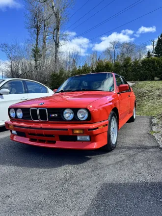 BMW M3 E30 rouge, trois quarts avant gauche, badge M3 sur l’aile.